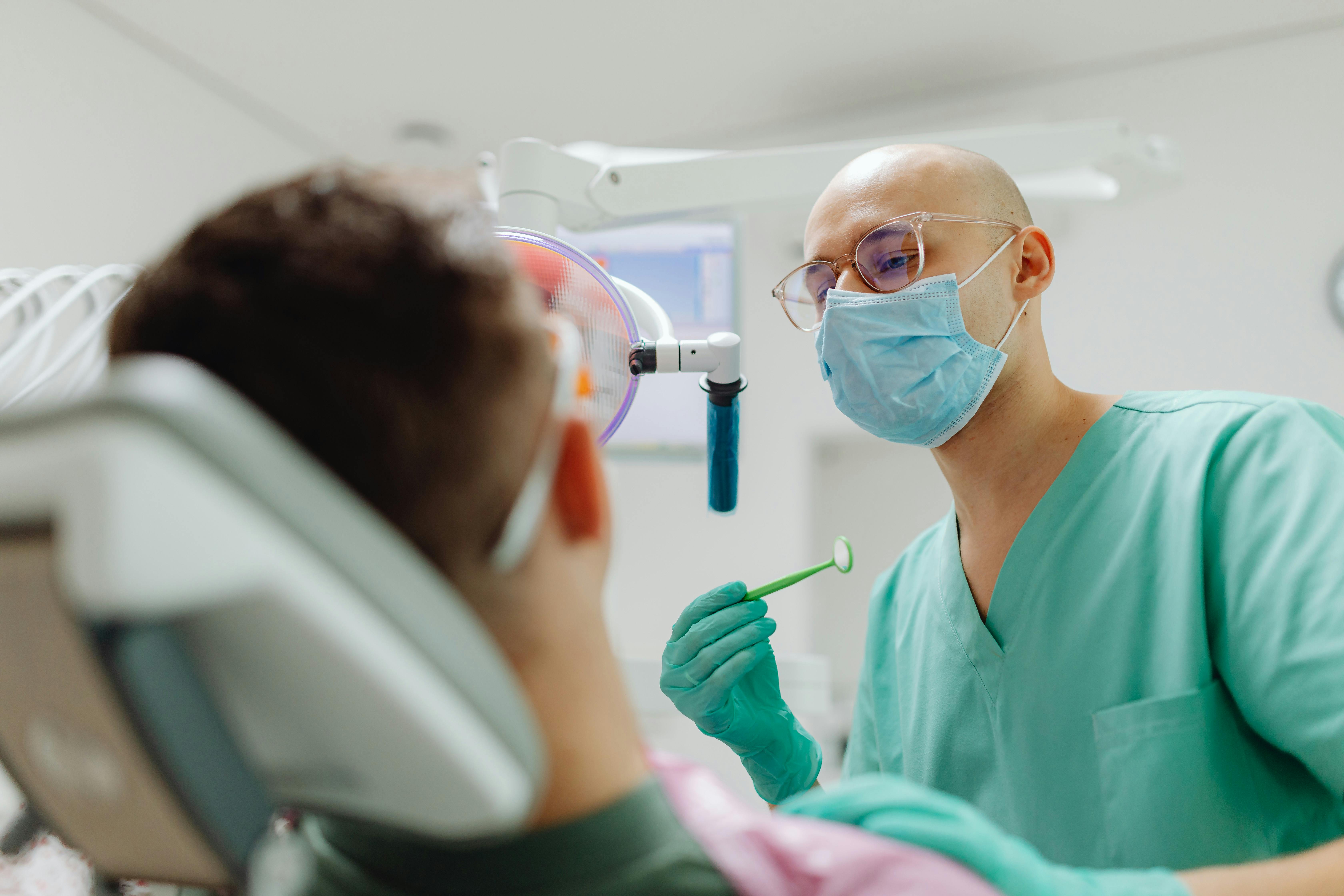 Un dentista con mascarilla y uniforme verde menta examina a un paciente desenfocado en una clínica dental.